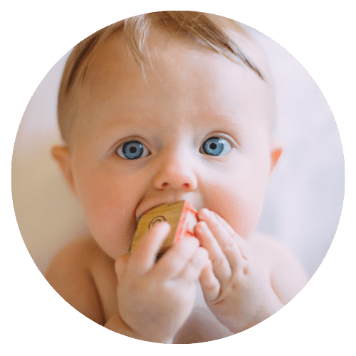 A baby with blue eyes holds a "baby" wooden block and brings it to their mouth.