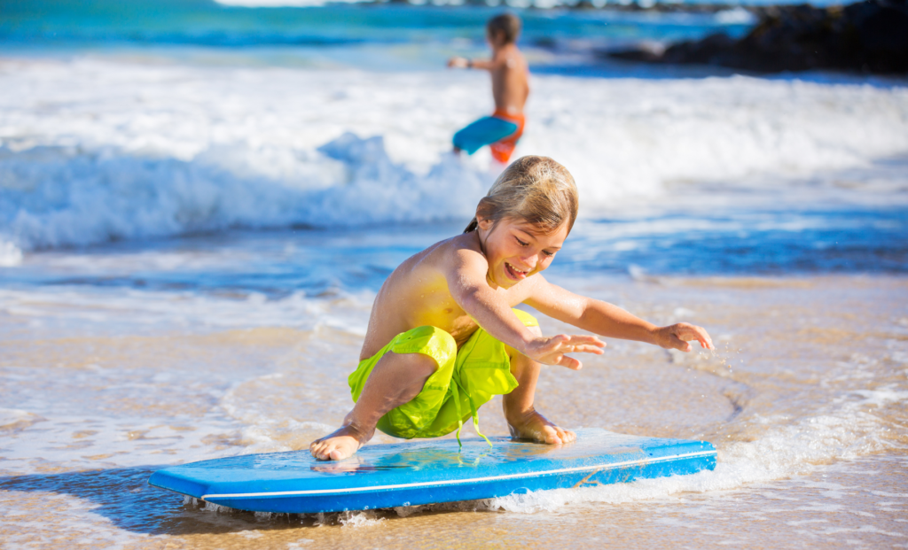 A child wearing green shorts rides a blue boogie board in ocean waves with another person nearby and a rocky background.